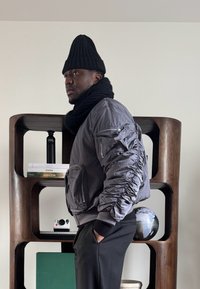 Homme portant un bonnet noir et une écharpe avec une veste grise, debout de profil, les mains dans les poches devant une étagère en bois avec des livres et un globe.