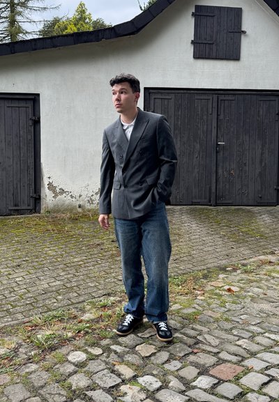 Blazer gris de doble botonadura con solapas rectas, combinado con jeans azul oscuro y zapatillas negras, con un fondo de un camino de piedra texturizado.