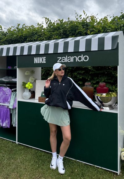 Mujer con un conjunto deportivo de Nike está de pie junto a un stand verde y blanco con los logotipos de Zalando y Nike, con estantes de ropa y bebidas visibles al fondo.