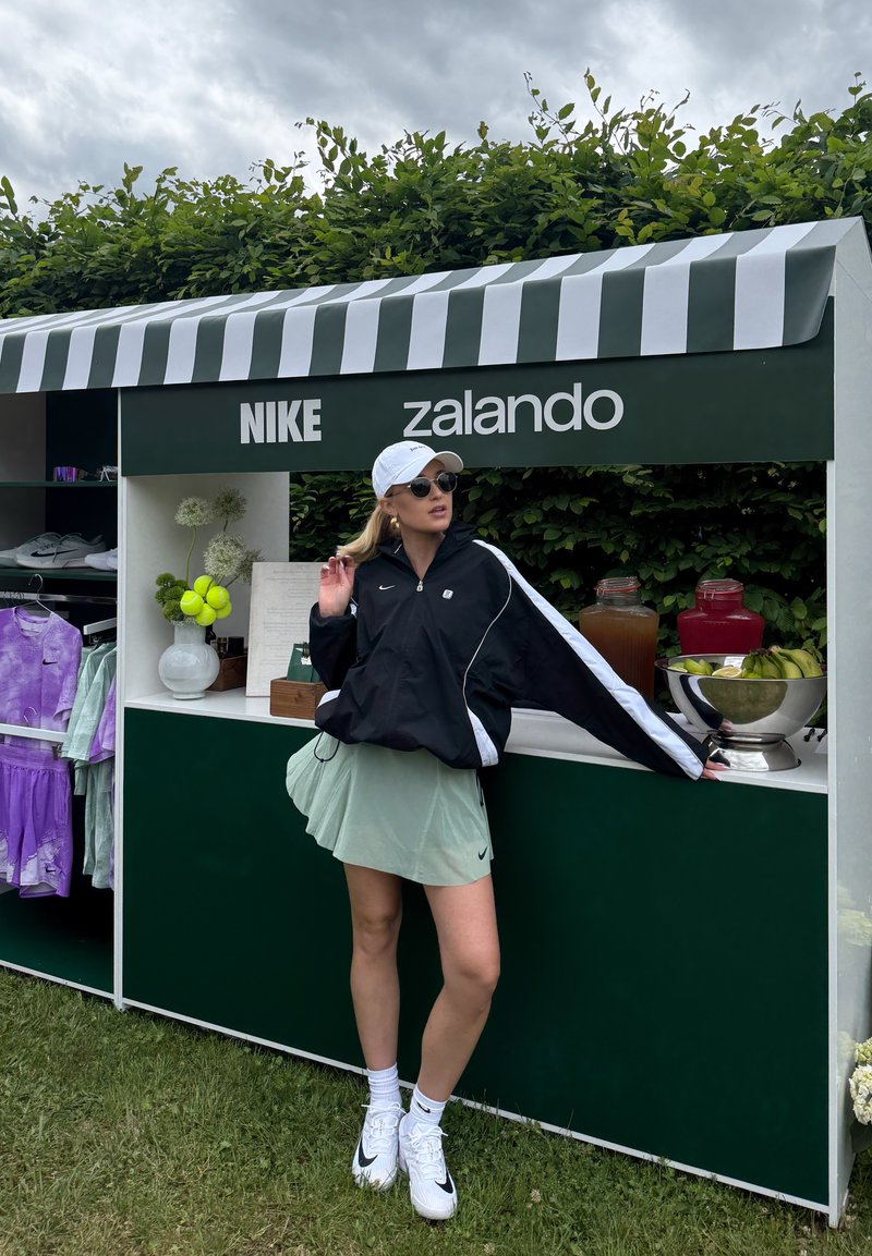 Femme en tenue sportive Nike debout à côté d’un stand vert et blanc avec les logos Zalando et Nike, des étagères de vêtements et de boissons visibles à l’arrière-plan.