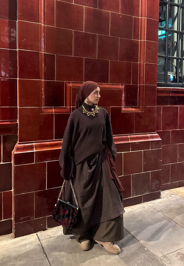 Frau mit braunem Hijab und geschichtetem braunem Outfit, goldener Halskette und Ohrringen, die eine gemusterte Handtasche hält und an einer roten gefliesten Wand steht.