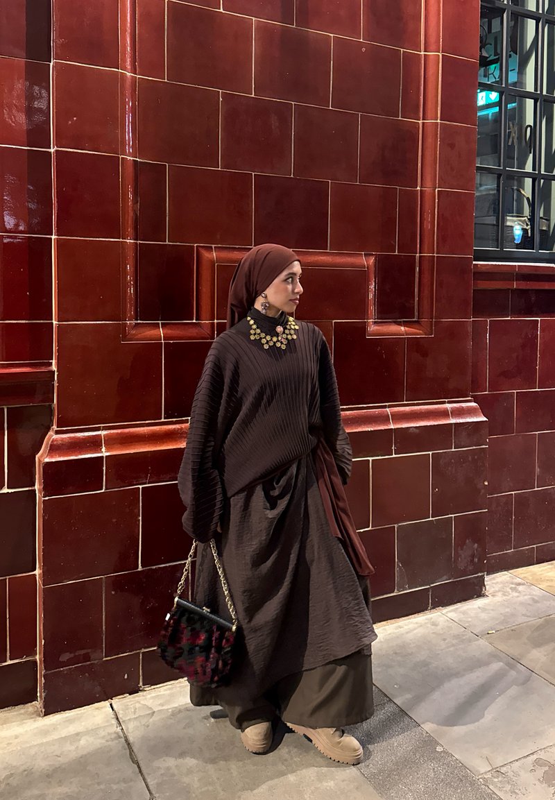 Femme portant un hijab marron et une tenue marron superposée avec un collier et des boucles d'oreilles en or, tenant un sac à main à motif, debout près d’un mur carrelé rouge.
