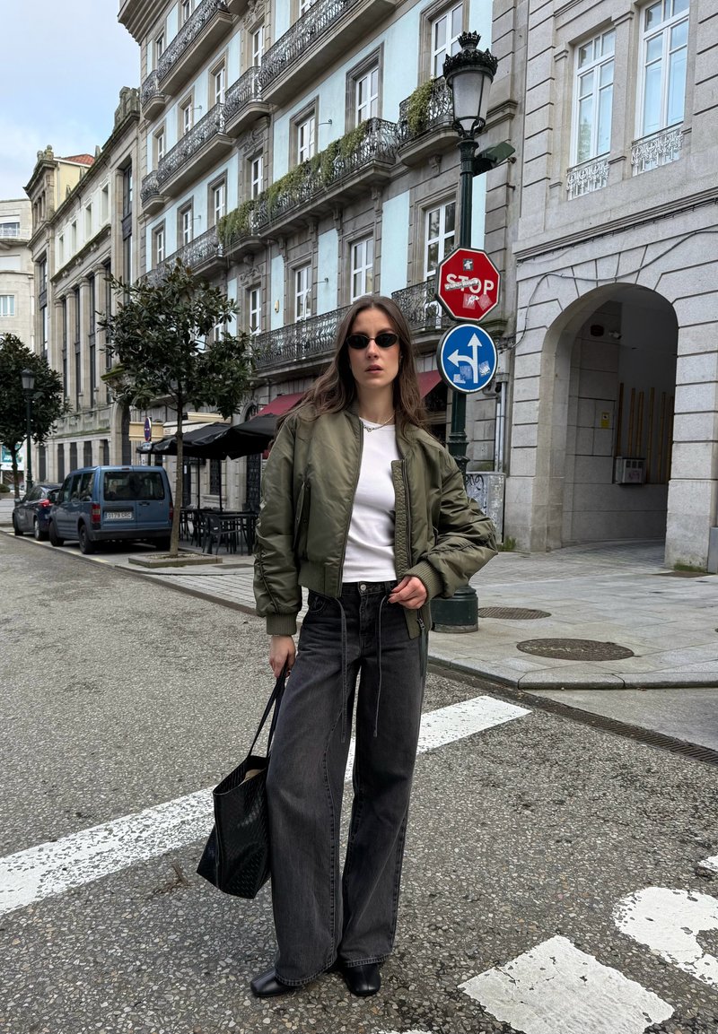 Femme portant des lunettes de soleil, une veste bomber verte, une chemise blanche, un jean noir large, tenant un sac cabas noir, debout dans une rue de la ville près de panneaux de signalisation.