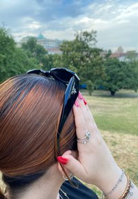 Sorte Prada-solbriller hviler på brunt hår, med en hånd placeret nær øret. Bemærkelsesværdig er den glinsende tekstur og det lagdelte frisure.