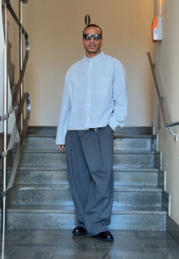 Man wearing sunglasses, a light striped shirt, and loose grey trousers standing on indoor stairs with metal railings.