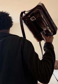 Personne aux cheveux bouclés, vêtue de noir, tenant un sacoche en cuir marron foncé poli sur l'épaule à l'intérieur.