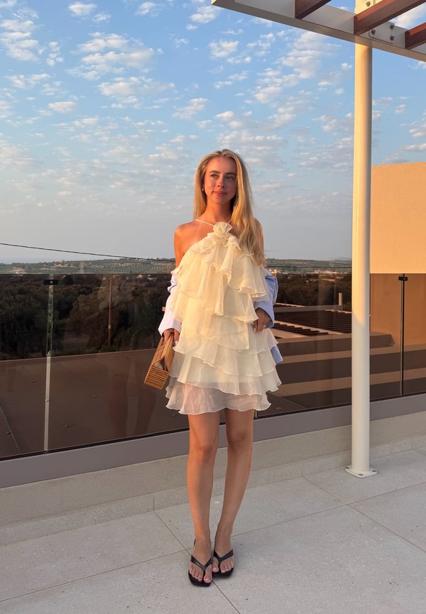 Joven mujer con un vestido blanco en capas y sandalias negras está en una terraza con un bolso de madera al atardecer.