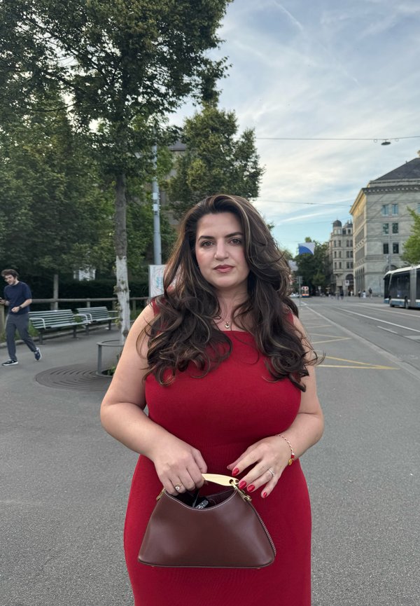 Frau mit langen dunklen Haaren trägt ein rotes Kleid, hält eine braune Handtasche und steht in einer Stadtstraße mit Bäumen und Gebäuden im Hintergrund.