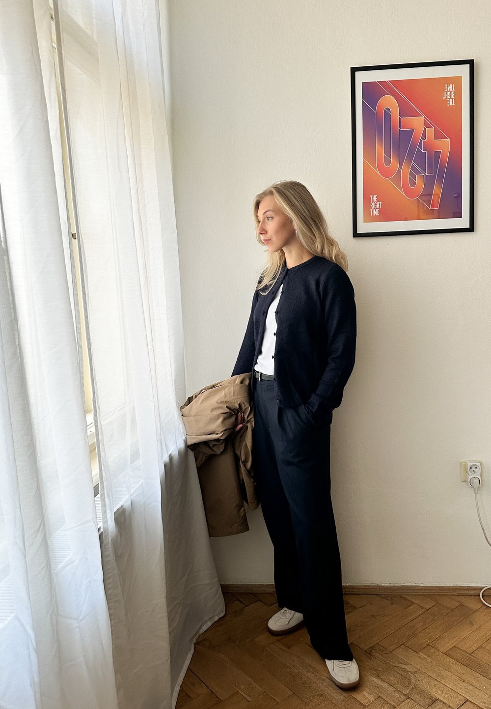 Un cardigan bleu marine sur une chemise blanche, un pantalon noir et des baskets blanches. Tenant un manteau beige clair. Lumière naturelle filtrée par des rideaux légers.