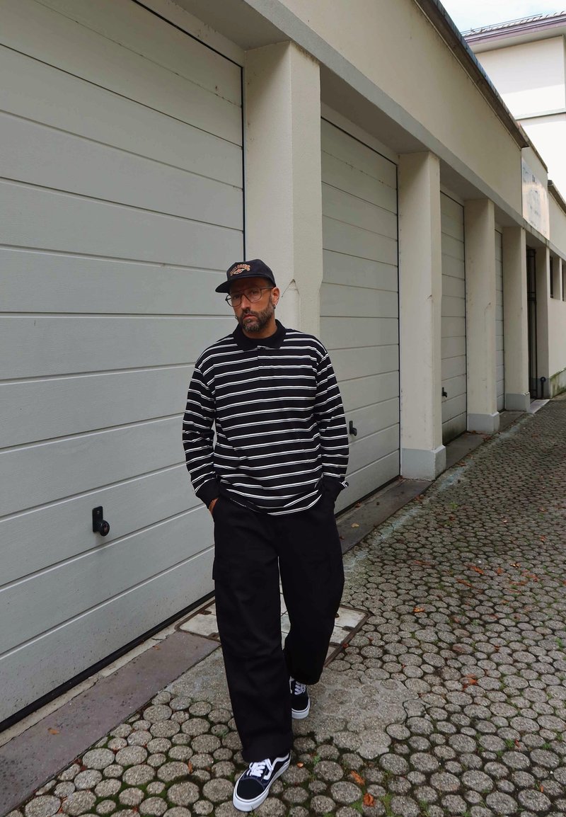 Uomo che indossa una camicia a maniche lunghe a righe bianche e nere, pantaloni neri, berretto nero e occhiali, mentre passa davanti a porte del garage chiare e chiuse.