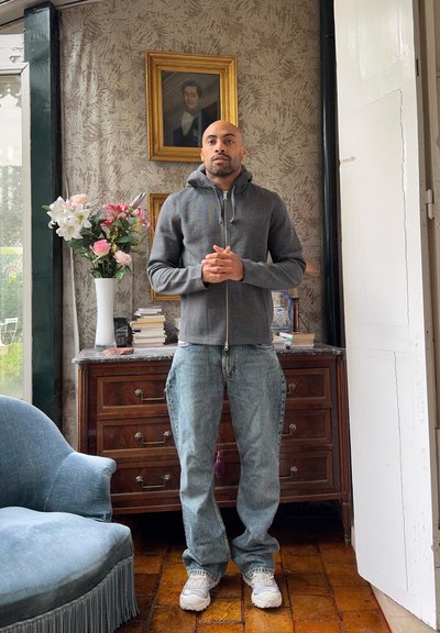 Sudadera gris con cremallera, vaqueros azules y zapatillas blancas, llevadas por una persona que está de pie en una habitación con un jarrón de flores, una cómoda de madera y papel pintado.