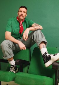 Homem em uma t-shirt verde texturizada e bandana vermelha senta-se em cadeiras verdes. Calças cinzentas e ténis de cano alto pretos completam o visual.