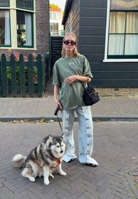 Pārmēri olīvu zaļš T-krekls, gaiši zili pogājami platā kāju bikses, melna somiņa, tumši saulesbrilles; pūkains suns ar pavadu ielās.