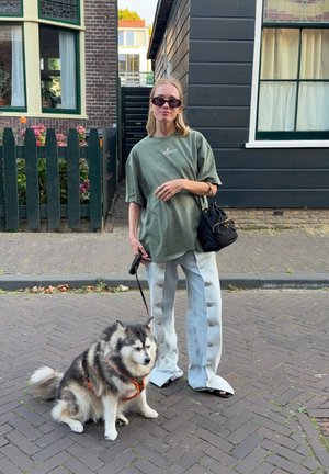 Camiseta de oliva de gran tamaño, pantalones anchos de botones en azul claro, bolso negro, gafas de sol oscuras; un perro esponjoso con correa en un entorno urbano.