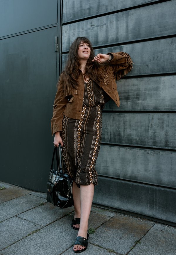 Femme aux longs cheveux portant une robe à motifs et une veste marron à franges, debout contre un mur gris, tenant un sac à main noir et souriant.