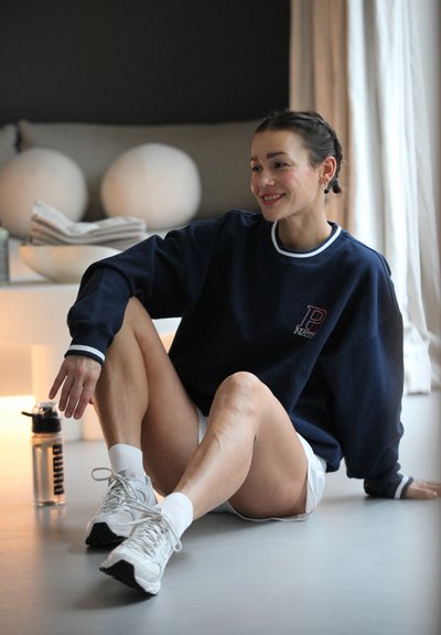 Sudadera navy con detalles en blanco, corte relajado, combinada con shorts blancos y zapatillas blancas. Botella transparente al lado. Fondo neutro.