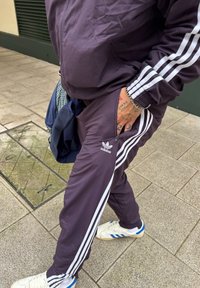 Survêtement Adidas violet avec trois bandes blanches, poches zippées, et sneakers blancs avec des accents bleus sur les côtés.