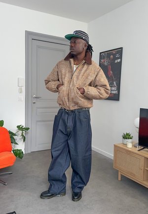 Hombre negro con chaqueta a cuadros, pantalones anchos de mezclilla oscura, gorra a rayas, de pie en una habitación moderna con póster deportivo y plantas.
