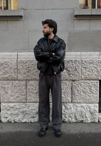 Hombre con barba y cabello oscuro está de pie con los brazos cruzados, vistiendo una chaqueta de cuero negra, pantalones grises y botas negras frente a una pared de piedra.