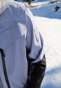 Veste de ski violet clair avec des accents noirs, équipée d'une fermeture éclair à l'avant, de grandes poches et d'un logo sur l'épaule. Arrière-plan enneigé visible.