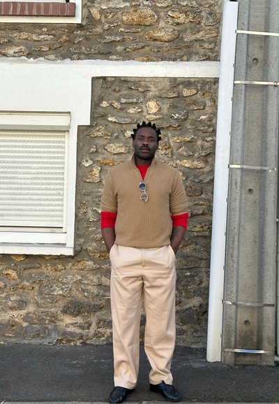 Hombre con trenzas lleva un suéter beige sobre una camisa roja de manga larga, pantalones beige, zapatos negros y gafas de sol colgadas del suéter, de pie junto a un muro de piedra.