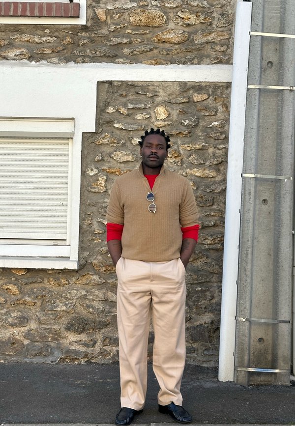 Man met gevlochten haar draagt een beige trui over een rood shirt met lange mouwen, crèmekleurige broek, zwarte schoenen en een zonnebril die aan de trui hangt, staand bij een stenen muur.