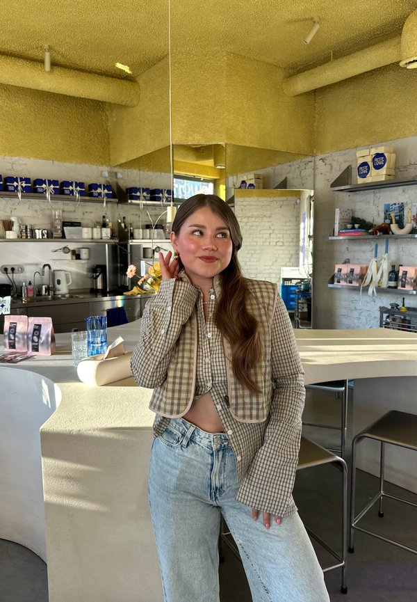 Jeune femme en veste à carreaux décontractée et jean posant à l'intérieur d'un café moderne avec des comptoirs blancs, des étagères et un plafond texturé jaune.