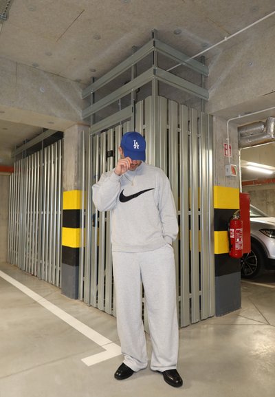 Sudadera gris con el logo negro de Nike y pantalones de chándal a juego, llevados con una gorra de béisbol azul de LA y zapatos negros, en un aparcamiento.
