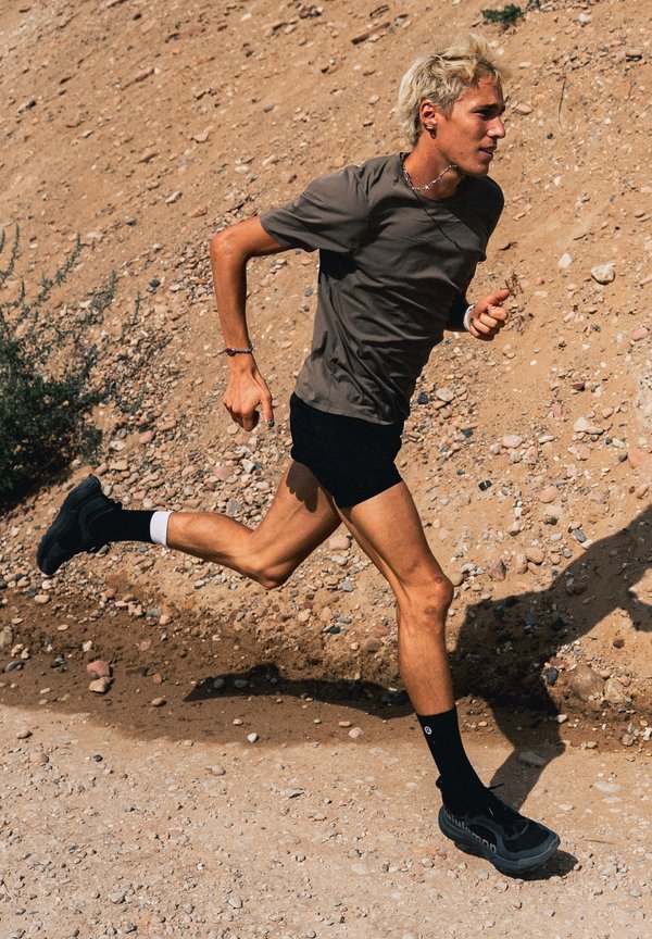 Joven con cabello rubio corriendo por un sendero rocoso de tierra, vistiendo una camiseta gris, pantalones cortos negros, calcetines negros y zapatillas negras para correr.