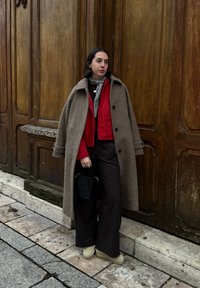 Femme portant un long manteau marron sur une veste rouge, un pantalon large noir, des chaussures crème, tenant un petit sac à main noir, debout près de portes en bois.