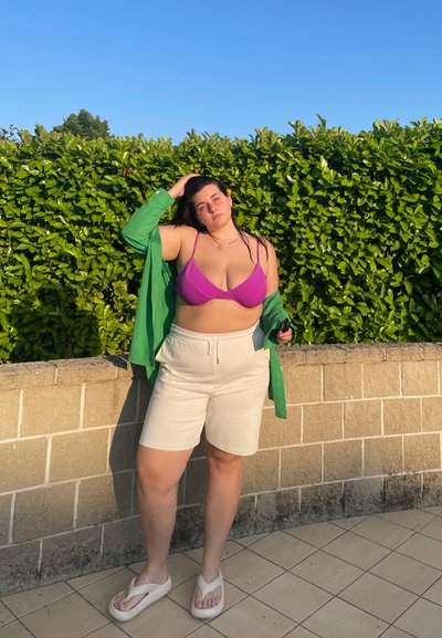 Sostenedor de color púrpura con canalé, camisa verde de botones, pantalones cortos beige y chanclas de color claro. El fondo presenta un seto verde y una pared de piedra.