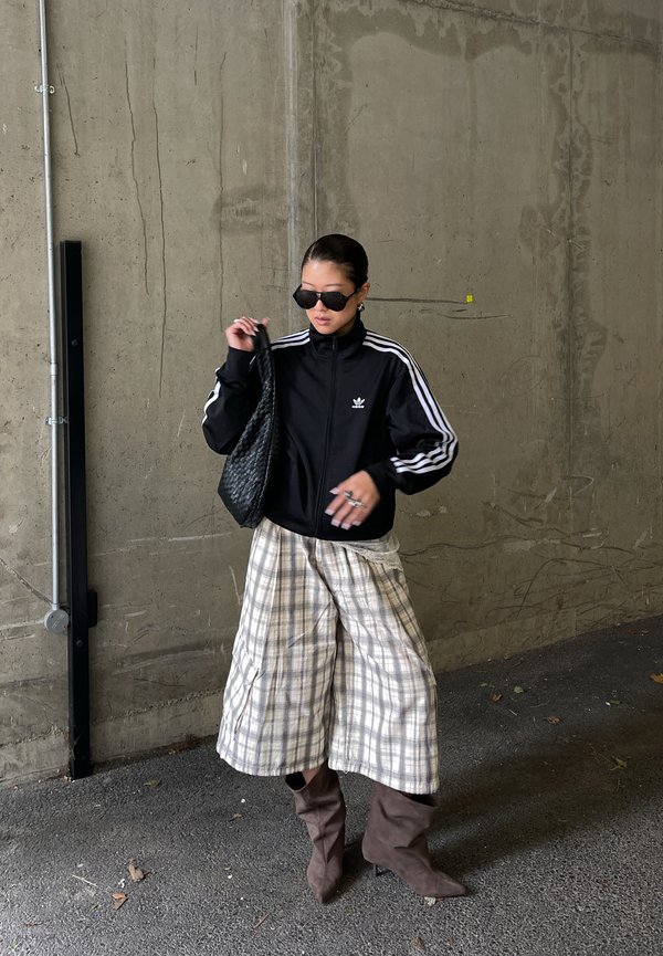 Persoană purtând o jachetă sport Adidas neagră, pantaloni largi în carouri, cizme maro, ochelari de soare negri, ținând o geantă neagră țesută, stând în picioare lângă un perete din beton.