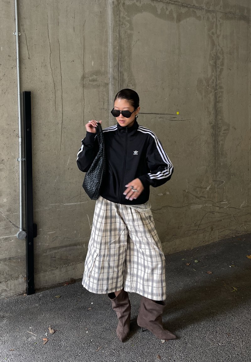 Άτομο που φοράει μαύρο αθλητικό μπουφάν Adidas, καρό φαρδιά παντελόνια, καφέ μπότες, μαύρα γυαλιά ηλίου, κρατάει μαύρη πλεκτή τσάντα, στέκεται μπροστά σε τσιμεντένιο τοίχο.