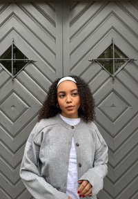 Cardigan en tricot gris avec boutonnière, poignets côtelés, porté sur un T-shirt blanc, devant une porte texturée en chevron gris foncé.