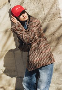 Femme portant une casquette rouge avec le texte "Darling", un blazer à carreaux et un jean bleu, appuyée contre un mur de pierre texturé au soleil.
