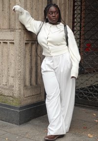Cardigan en tricot blanc avec un devant boutonné, associé à un pantalon blanc ample et des bottines marron, le tout devant un mur en pierre texturé.
