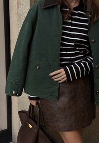 Persoon in een groene jas, zwart-wit gestreepte langemouwen shirt, bruine leren rok, met een grote bruine handtas.
