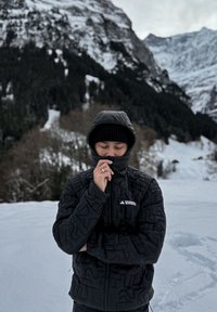 Personne portant une veste noire à capuche et un bonnet, zippée jusqu'au menton, debout dans la neige avec des montagnes enneigées et des arbres en arrière-plan.