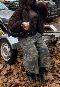 Femme portant une veste en fourrure marron et un pantalon camouflage, tenant un café glacé, debout sur des feuilles d'automne à côté d'une remorque de voiture dans une rue de la ville.