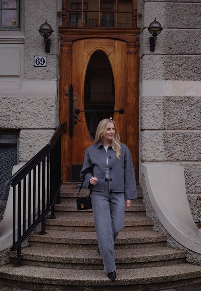 Mujer vestida con chaqueta gris y jeans, llevando un bolso negro, bajando escaleras de piedra en curva frente a una puerta de madera con el número de casa 69.