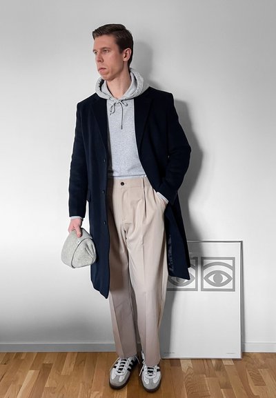 Hombre con abrigo negro, sudadera con capucha gris, pantalones beige, zapatillas blancas, sosteniendo una gorra gris, de pie junto a una obra de arte enmarcada con ilustraciones de ojos.