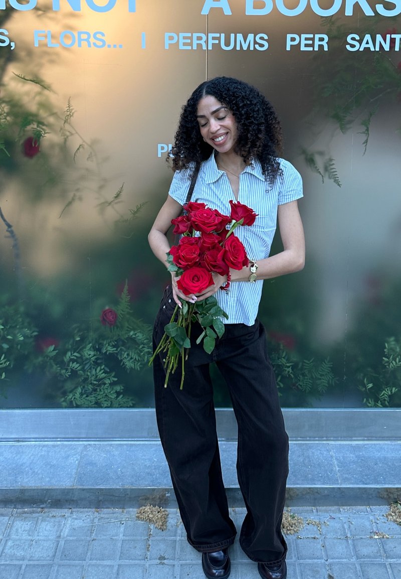 Femme aux cheveux bouclés portant une chemise rayée et un pantalon noir, souriante, tenant un bouquet de roses rouges devant un mur de verre avec du texte.