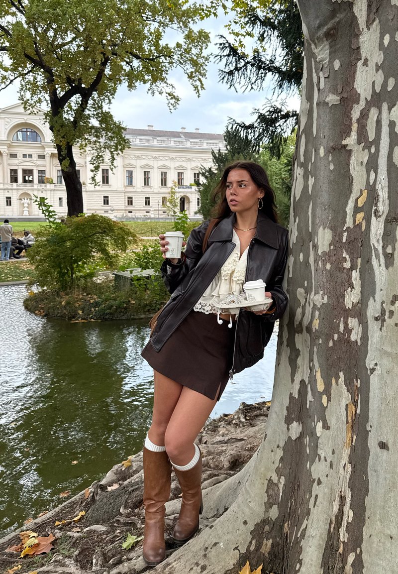 Mulher vestindo casaco de couro castanho e botas está junto a uma árvore perto de um lago, segurando copos de café e uma bandeja, com um edifício histórico ao fundo.
