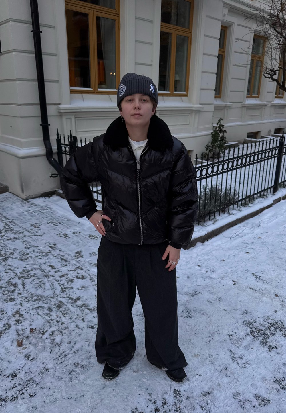 Doudoune noire avec col en fausse fourrure, fermeture éclair, associée à un pantalon large et foncé. Environnement comprenant des pavés recouverts de neige.