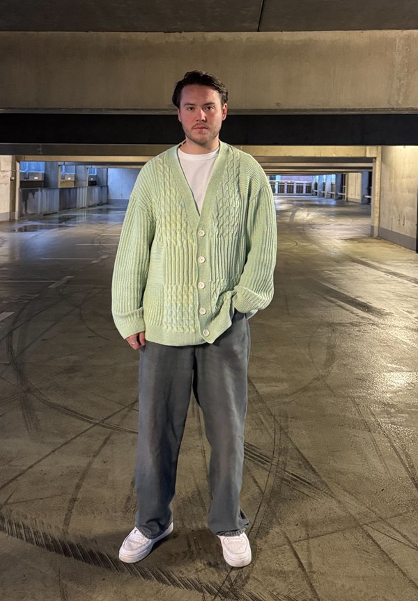 Hellgrüne Strickjacke mit einem Zopfmuster, fünf Knöpfen, über einem weißen Hemd getragen, kombiniert mit locker sitzenden grauen Hosen und weißen Schuhen.