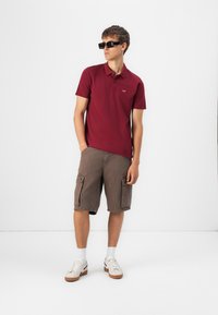 Burgundy polo shirt featuring a collar and logo, teamed with brown cargo shorts. The model is wearing black sunglasses and white trainers with beige details.