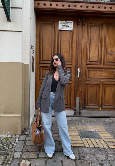 Blazer gris a rayas, top negro y jeans de pierna ancha azul claro. Bolsa de mano marrón, gafas de sol redondas. Escenario al aire libre con un fondo de puerta de madera.
