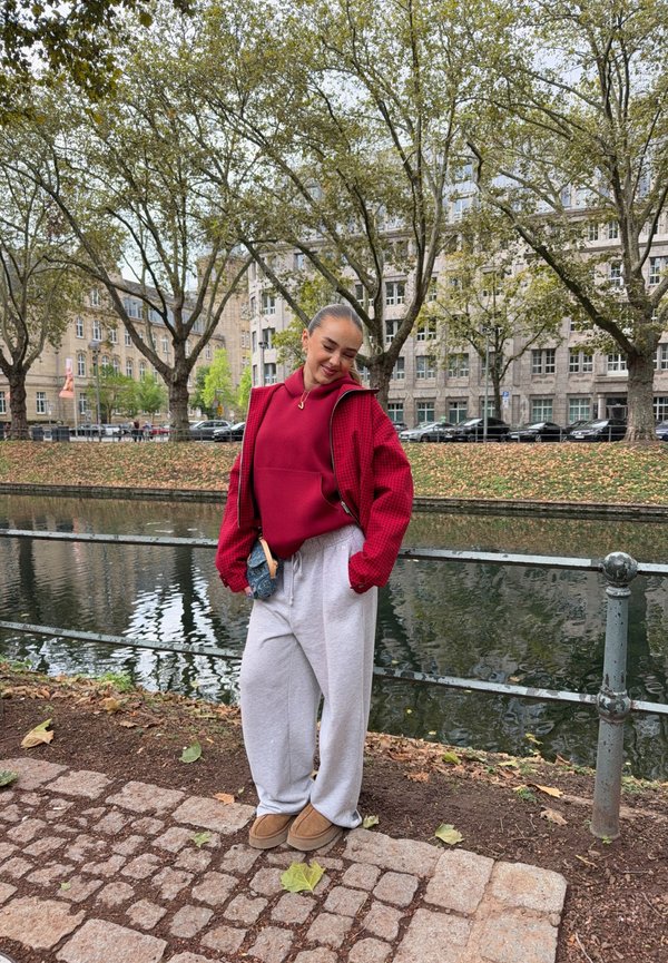 Joven mujer con chaqueta roja y pantalones grises de pie cerca de un canal con árboles y edificios al fondo en un día nublado.