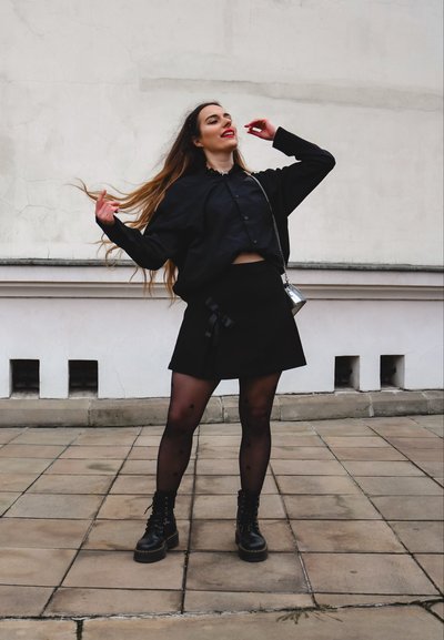Camisa corta negra, falda negra de cintura alta con detalle de cinta, medias negras con estampado y botas de combate negras. Bolsa cruzada plateada.