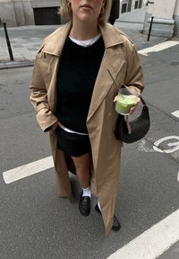 Trench-coat beige par-dessus un pull noir et une jupe noire. Chaussettes blanches avec des mocassins noirs. Tenant une boisson verte dans un verre transparent.